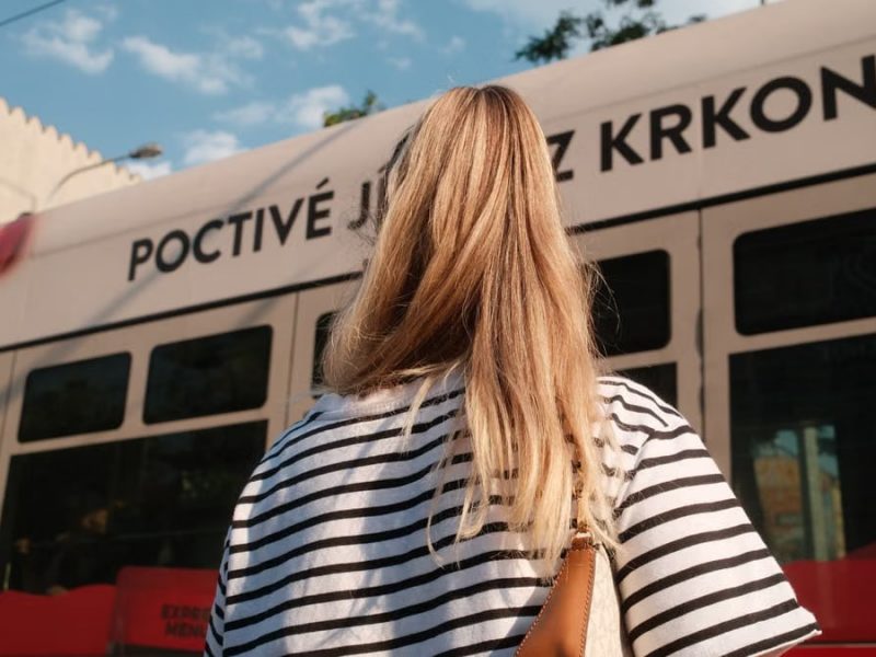 A woman with blonde hair in a striped shirt stands near a city tram on a sunny day.
